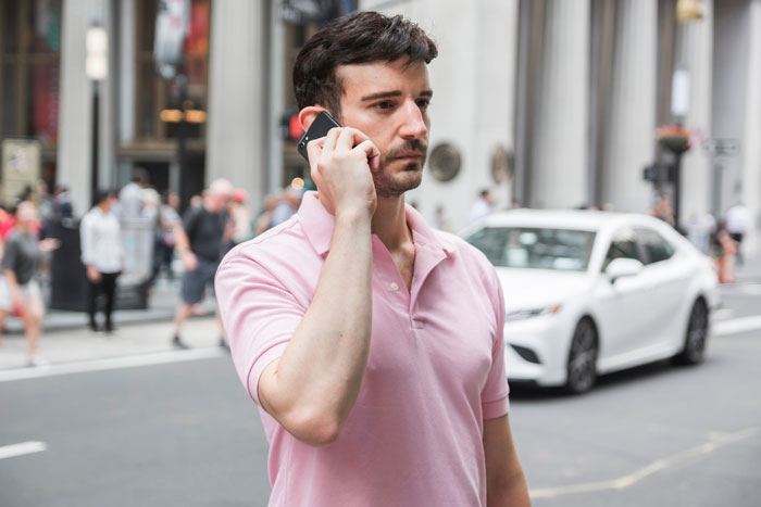 Man in pink shirt on phone outdoors in city, appearing concerned about surprise family visit during surgery plans. Man in pink shirt on phone outdoors in city, appearing concerned about surprise family visit during surgery plans.