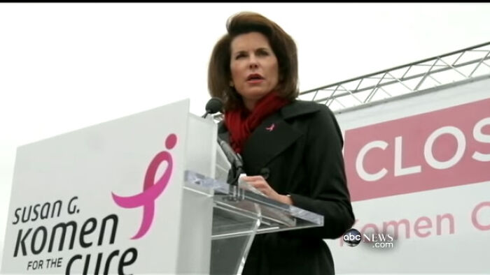 Woman speaking at a Susan G. Komen event, standing at a podium with a microphone and breast cancer awareness symbol.