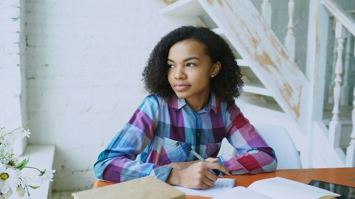 Young woman in a plaid shirt sitting at a desk, reflecting thoughtfully while writing about longest grudges held.