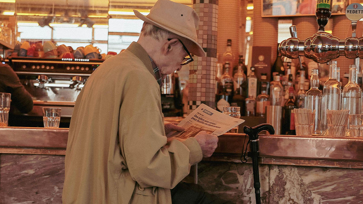 Elderly man sitting alone at a bar, reading a newspaper, capturing a painful table for one moment in a restaurant setting.