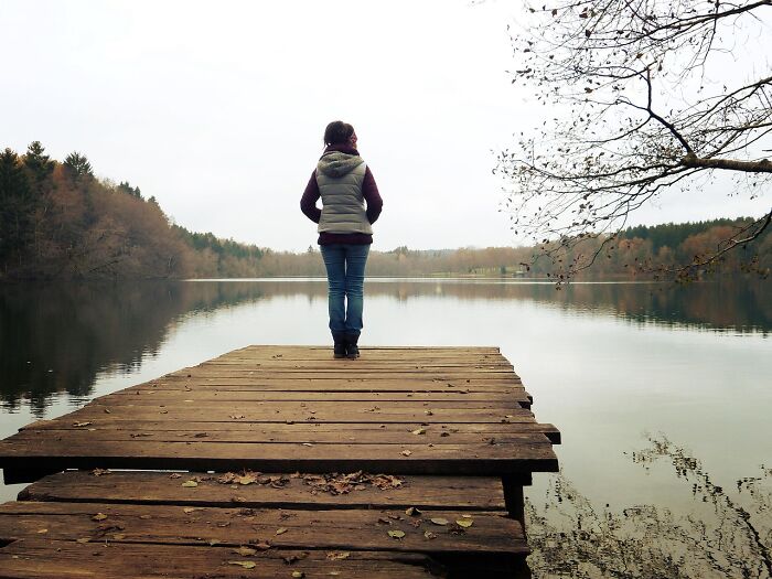 Woman standing alone on a wooden dock overlooking calm lake surrounded by autumn trees, reflecting unexpected symptoms.
