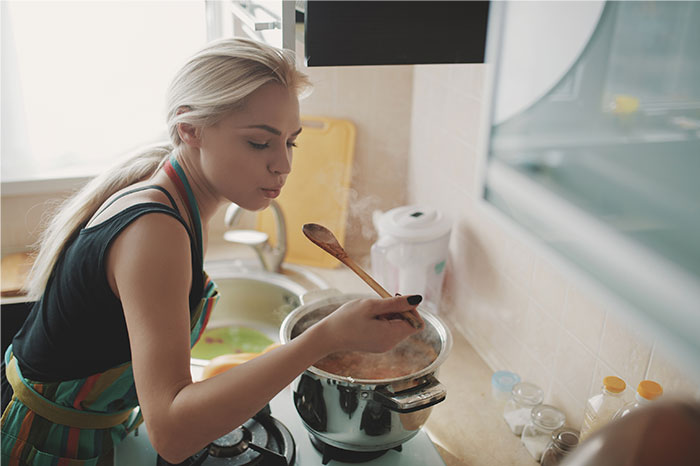 Woman in apron tasting food from a pot on stove, demonstrating kitchen hacks for cooking smarter with chicken.