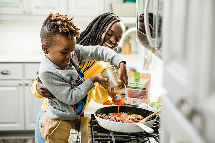 Child pouring sauce into a skillet while an adult helps, demonstrating cooking smarter kitchen cheats with I blow dry chicken method.