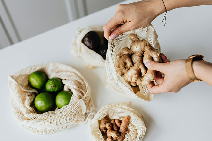 Hands holding fresh ginger over reusable mesh bags with limes and turmeric, illustrating smart kitchen storage hacks.