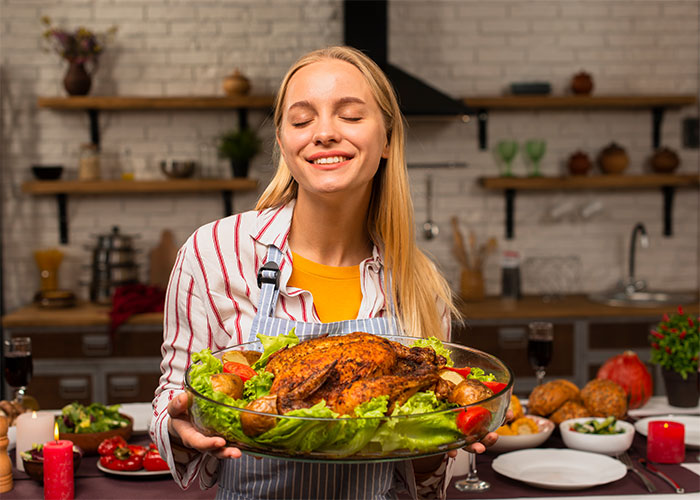 Young woman smiling and holding a roasted chicken surrounded by vegetables in a cozy kitchen setting.
