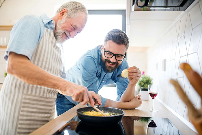 Older man blow drying chicken in pan while younger man tastes food in modern kitchen, showing cooking smarter kitchen cheats.