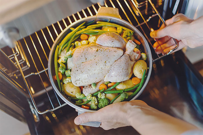 Person placing a whole seasoned chicken with vegetables into an oven, illustrating a smart cooking kitchen cheat.