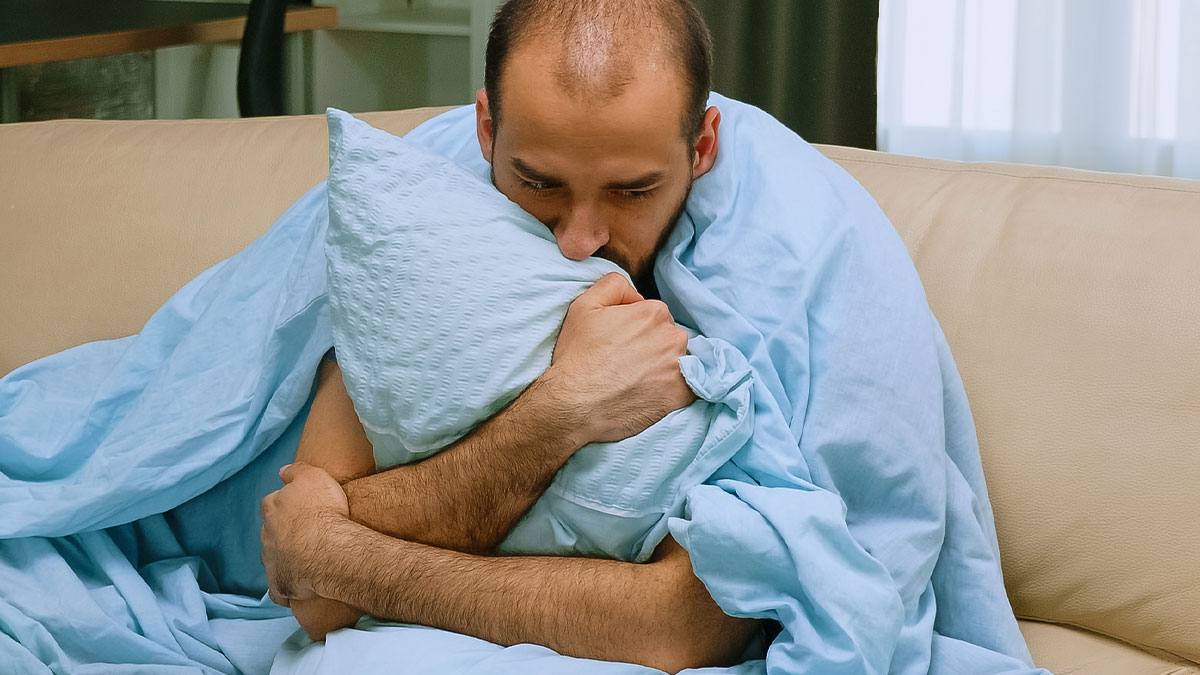 Worried dad wrapped in blue blanket hugging pillow on couch, illustrating dad in maternity wards stories.