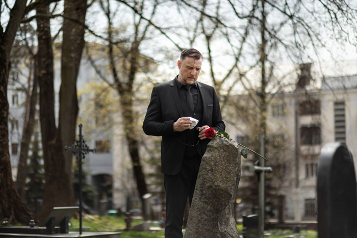 Man in black suit holding a red rose at a gravestone, reflecting on dying wife’s wishes about dating after loss. Man in black suit holding a red rose at a gravestone, reflecting on dying wife’s wishes about dating after loss.