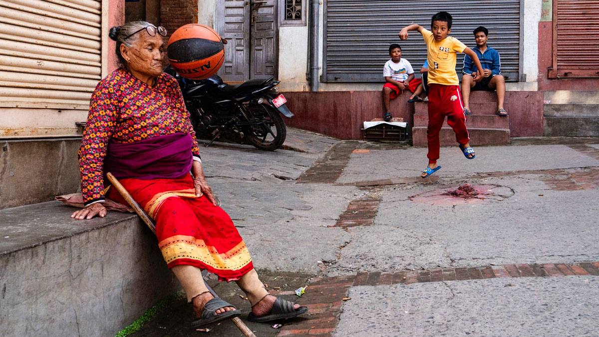 Elderly woman sitting on a street curb while children play basketball nearby in a vibrant street photography scene.