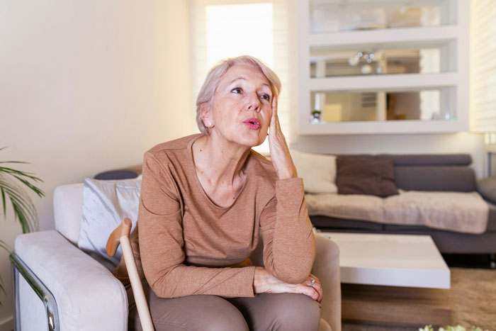 Older woman sitting on chair at home, looking thoughtful and stressed, relating to single mom plastic surgery support struggles. Older woman sitting on chair at home, looking thoughtful and stressed, relating to single mom plastic surgery support struggles.
