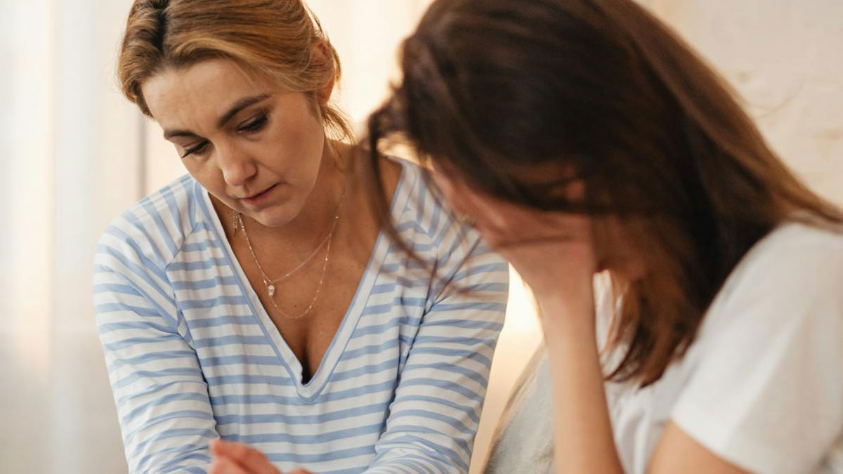 Two women sitting indoors, one looking upset while the other covers her face, capturing parental guilt-tripping tension.