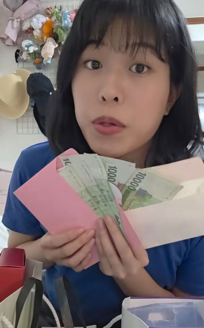 Woman holding cash gifts received while interviewing for jobs in Korea, sitting indoors with bags and plush toys in background Woman holding cash gifts received while interviewing for jobs in Korea, sitting indoors with bags and plush toys in background