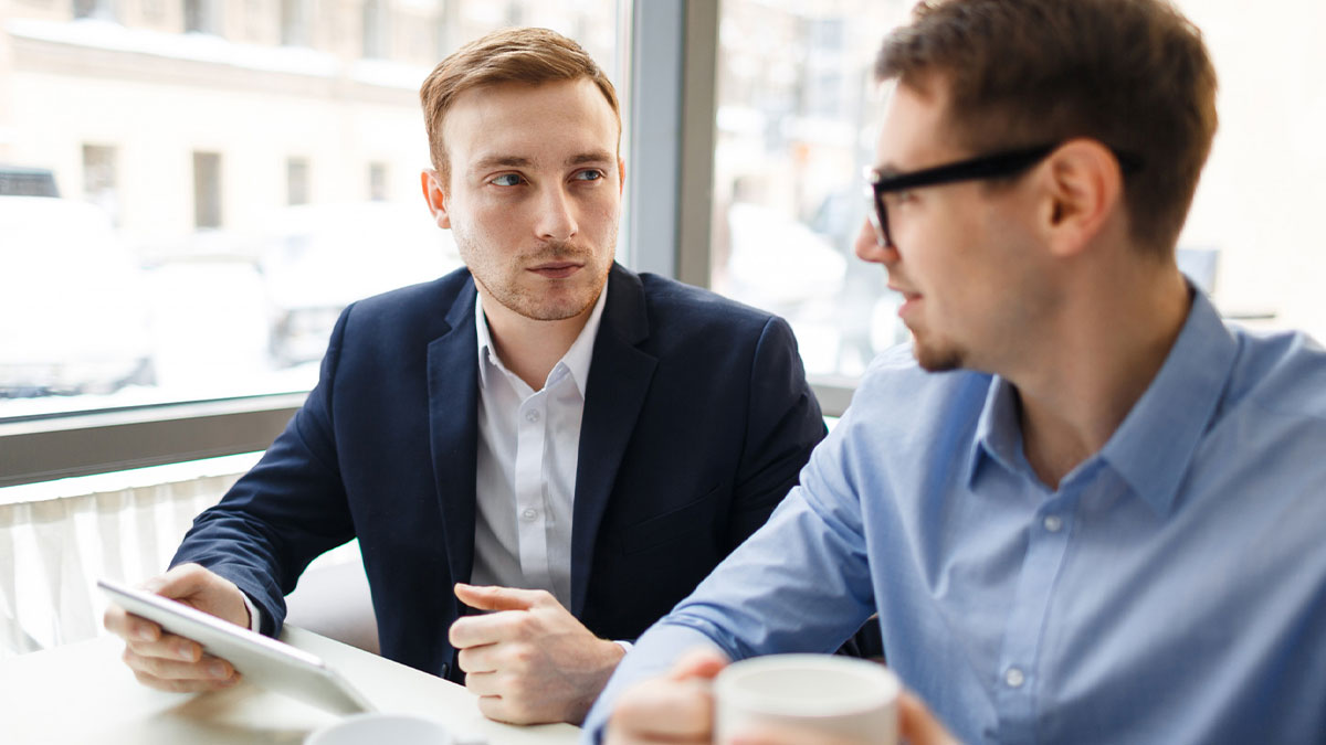 Two men at work having a quiet conversation, one holding a tablet, in an office with natural light.