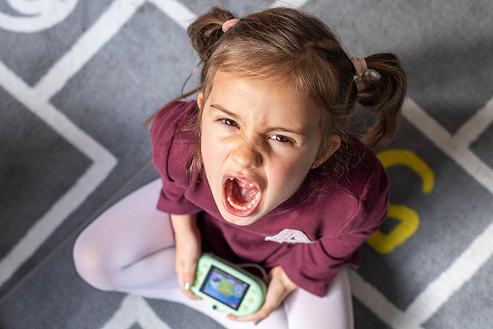 Young girl sitting on patterned carpet, angrily yelling while holding a handheld gaming device, babysitter moment captured.