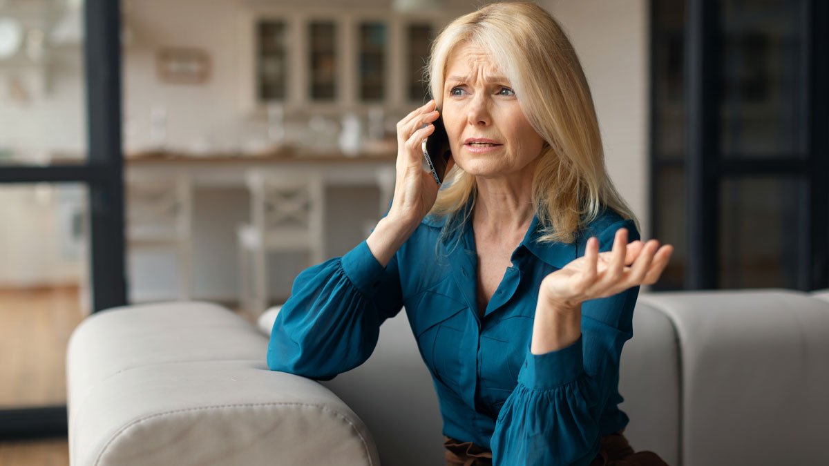 Woman in teal shirt sitting on couch, looking upset while talking on phone about neighbors from hell problems.