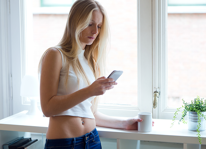 Young woman standing near window, looking at phone, reflecting on caregiver role preparation for disabled stepsister. Young woman standing near window, looking at phone, reflecting on caregiver role preparation for disabled stepsister.