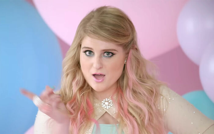 Meghan Trainor with blonde and pink hair, wearing a sparkling necklace, reacting with a surprised expression in a colorful setting.