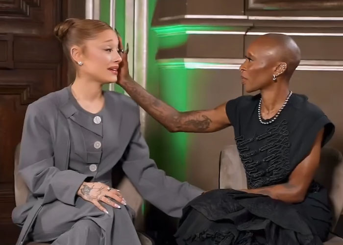 Ariana Grande and Cynthia Erivo seated and interacting during a candid moment amid interview discussions. Ariana Grande and Cynthia Erivo seated and interacting during a candid moment amid interview discussions.