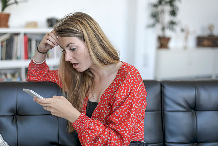 Woman in red blouse looking shocked at her phone, representing a mom receiving a surprising babysitting bill. Woman in red blouse looking shocked at her phone, representing a mom receiving a surprising babysitting bill.