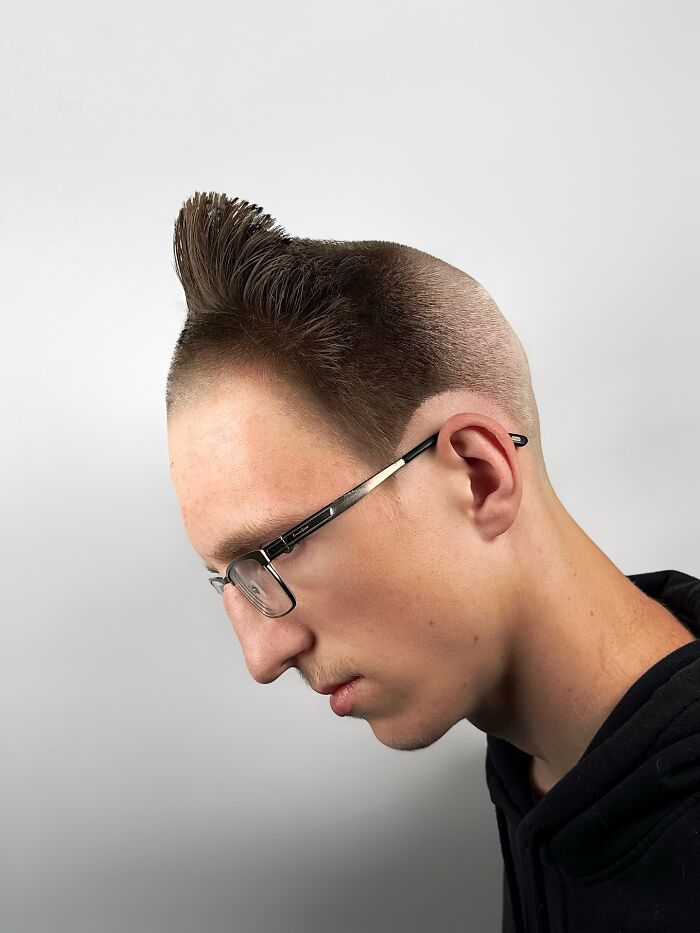 Young man with bold barber haircut styled upwards, wearing glasses and a black hoodie against a plain background