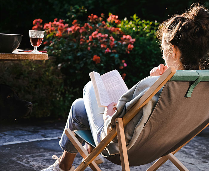 Woman reading romantasy book outdoors in a garden chair with flowers in the background on a sunny day