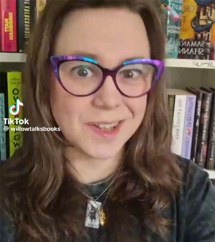 Woman with colorful glasses discussing romfantasy books in front of a bookshelf in a TikTok video.