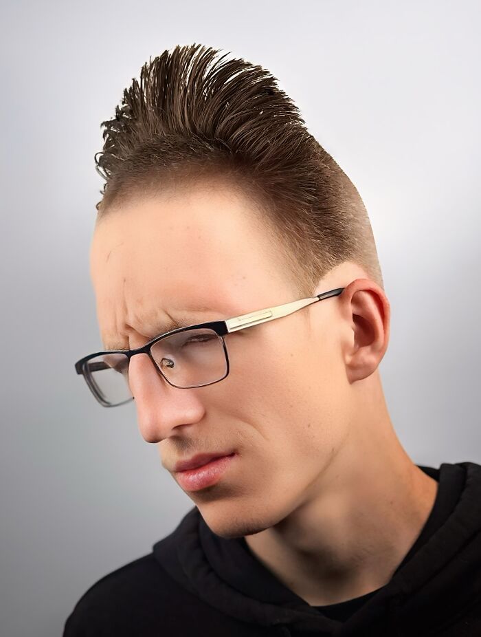 Young man with bold barber haircut styled upwards, wearing glasses and a black hoodie against a plain background