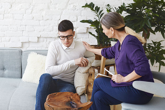 A concerned woman talks to a young man with autism on a couch, showing support and understanding.
