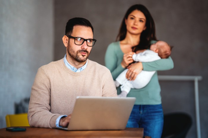 Man prioritizes work at laptop while wife stands behind holding baby, showing emotional distance in family.