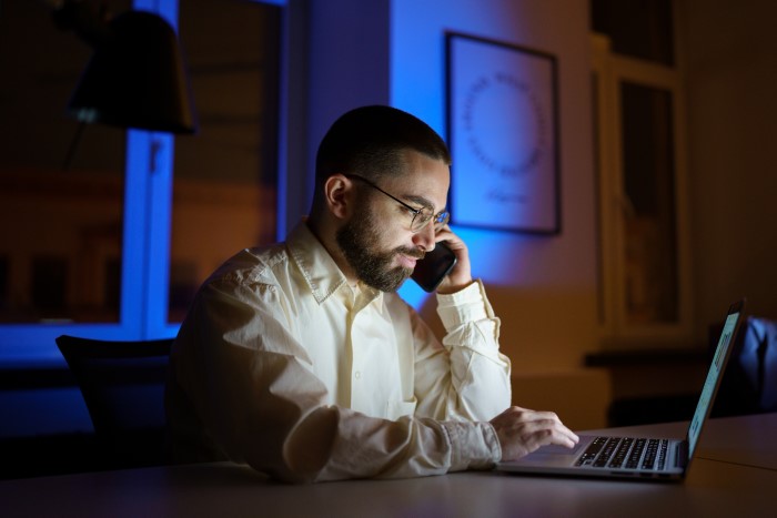 Man prioritizes work late at night, talking on phone and using laptop, ignoring family life and personal time.