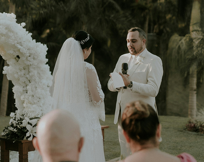 Groom reading wedding vows with microphone at outdoor ceremony, bride standing nearby in lace dress and veil. Groom reading wedding vows with microphone at outdoor ceremony, bride standing nearby in lace dress and veil.