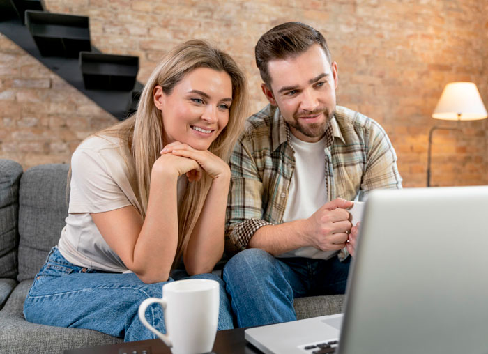 Couple looking at laptop together on couch, discussing vacation plans while dealing with MIL joining their trip. Couple looking at laptop together on couch, discussing vacation plans while dealing with MIL joining their trip.
