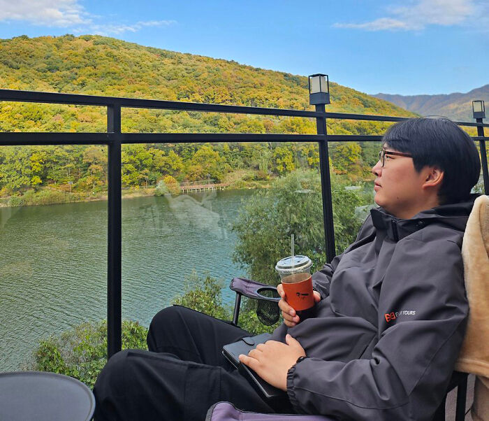 A man from South Korea wearing glasses and a jacket, sitting outdoors by a lake with autumn foliage. A man from South Korea wearing glasses and a jacket, sitting outdoors by a lake with autumn foliage.