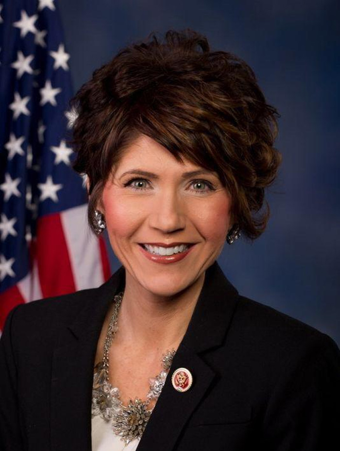 Kristi Noem smiling in a formal portrait with short styled hair and American flag in the background.