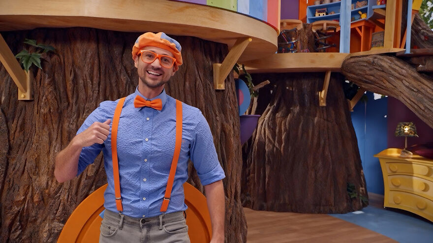 A Blippi actor smiling indoors wearing a blue shirt, orange bow tie, suspenders, and hat in a colorful set background. A Blippi actor smiling indoors wearing a blue shirt, orange bow tie, suspenders, and hat in a colorful set background.