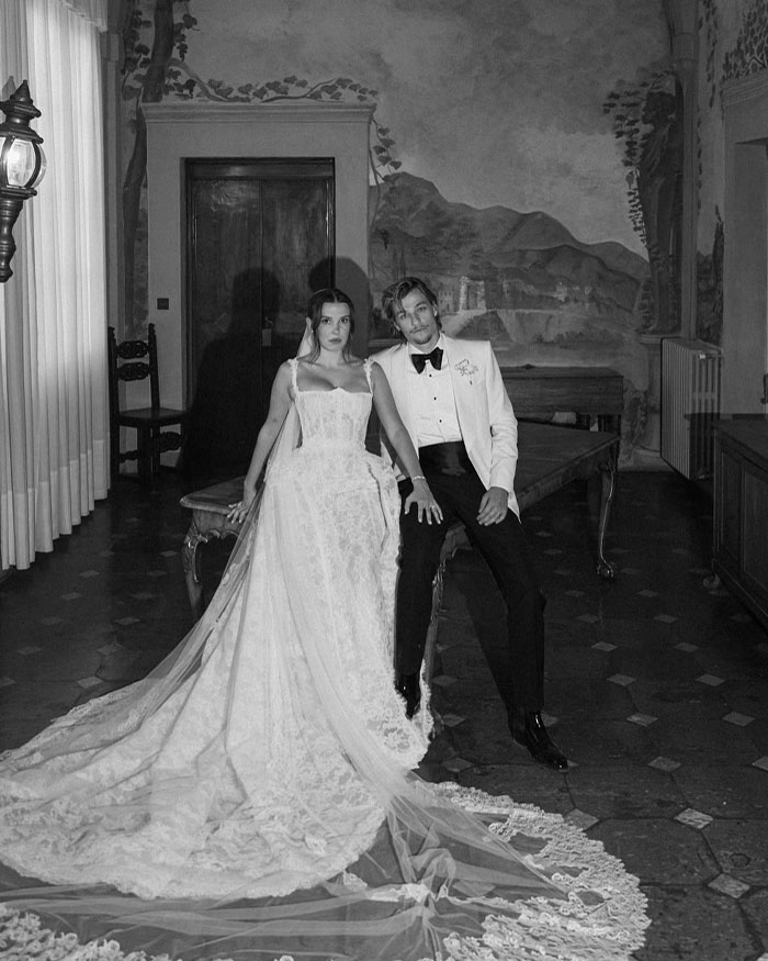 Black and white photo of a couple in formal attire, with Millie Bobby Brown wearing a long lace wedding dress indoors. Black and white photo of a couple in formal attire, with Millie Bobby Brown wearing a long lace wedding dress indoors.