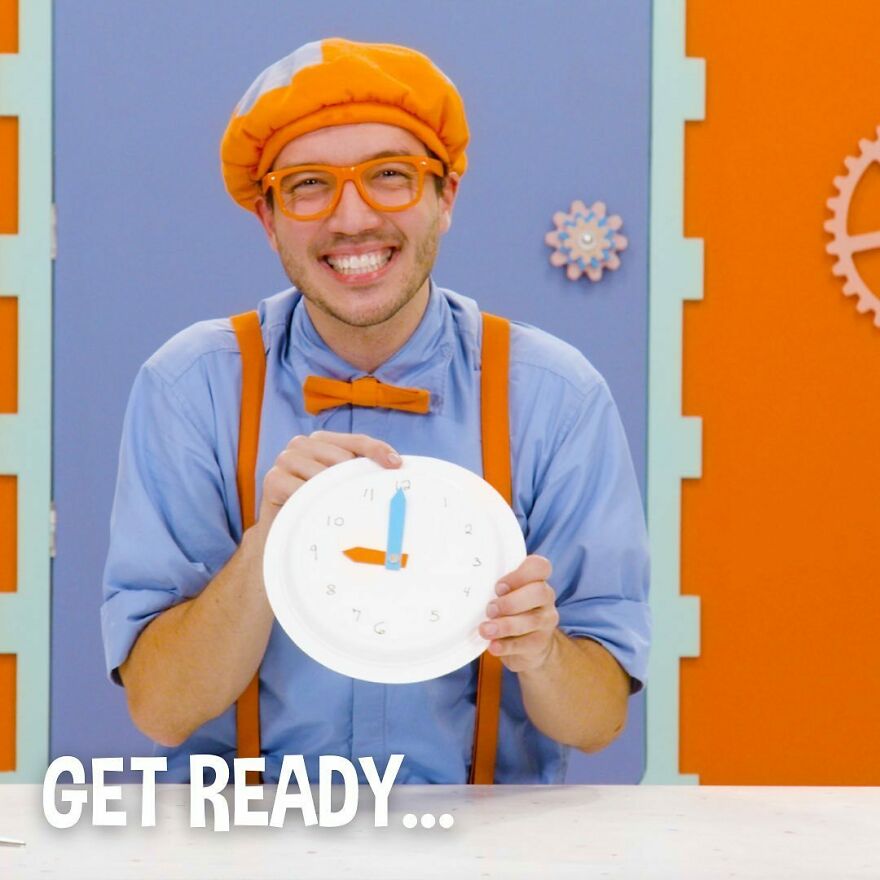 Smiling actor dressed as Blippi wearing orange bow tie, suspenders, and glasses, holding a paper clock in colorful setting. Smiling actor dressed as Blippi wearing orange bow tie, suspenders, and glasses, holding a paper clock in colorful setting.
