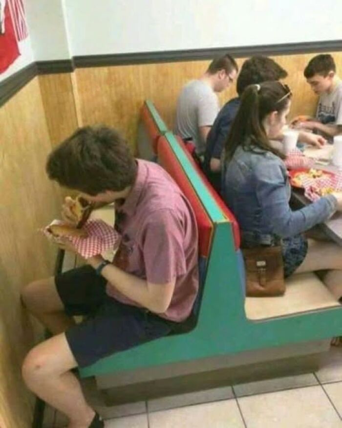 Young man sitting in a diner booth eating a burger, with other people enjoying food at the adjacent table of pics that go hard.