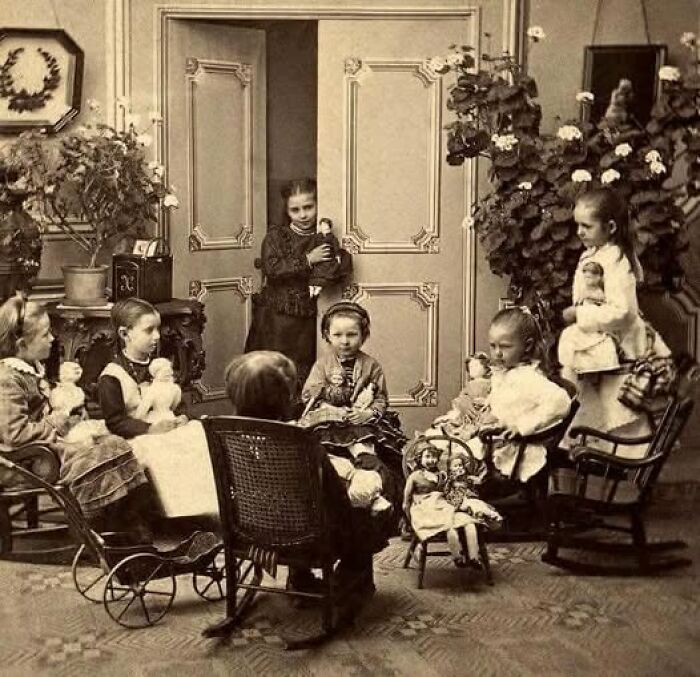 Group of Victorian-Edwardian children playing with dolls in a detailed vintage room with ornate walls and plants.