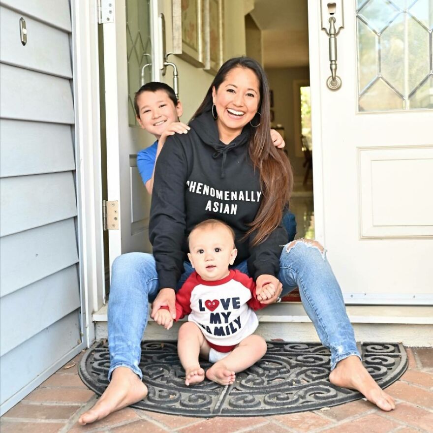 Smiling Asian woman with two children sitting on a doorstep wearing a hoodie and shirt promoting Asian pride and family love.