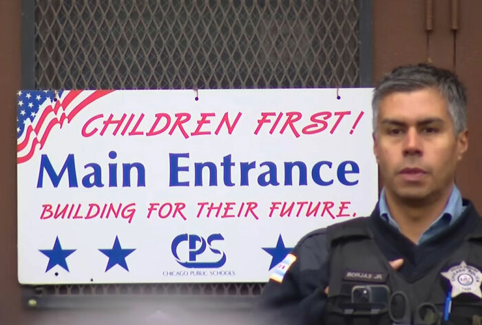 Police officer standing near Chicago Public Schools main entrance sign with children first message visible. Police officer standing near Chicago Public Schools main entrance sign with children first message visible.