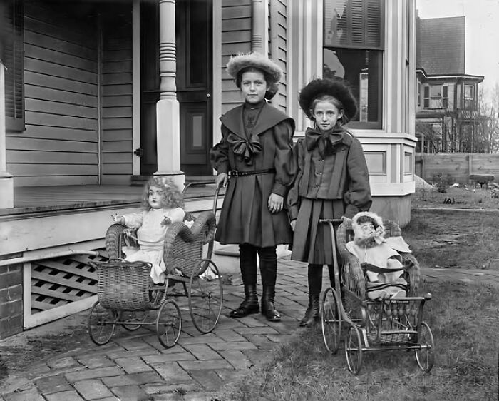 Two young girls in Victorian-Edwardian dresses standing outside with vintage dolls in wicker prams on a brick path.