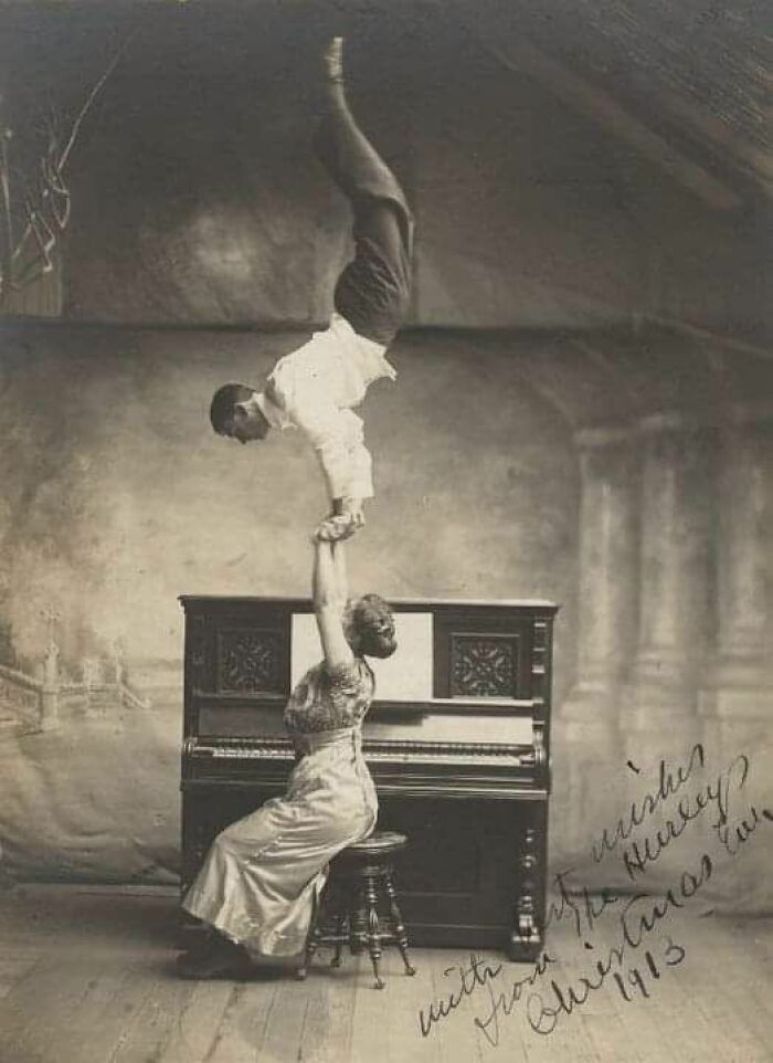 Woman in Victorian-Edwardian pictures holding man performing handstand on her hand near an old piano.