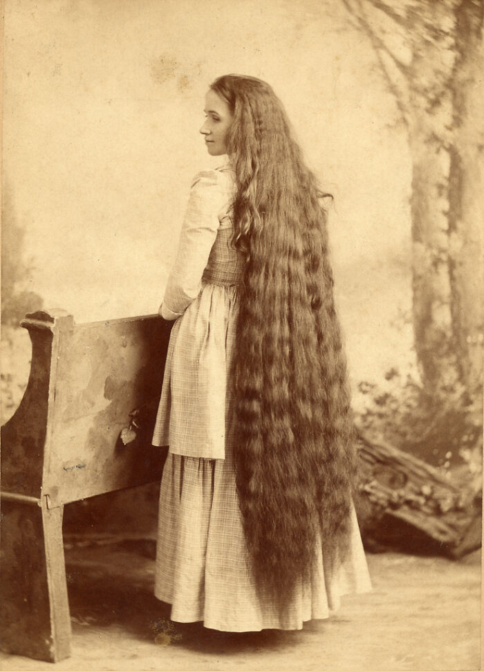 Young woman with extremely long hair wearing Victorian-style dress, posing indoors near wooden bench in Victorian-Edwardian pictures.