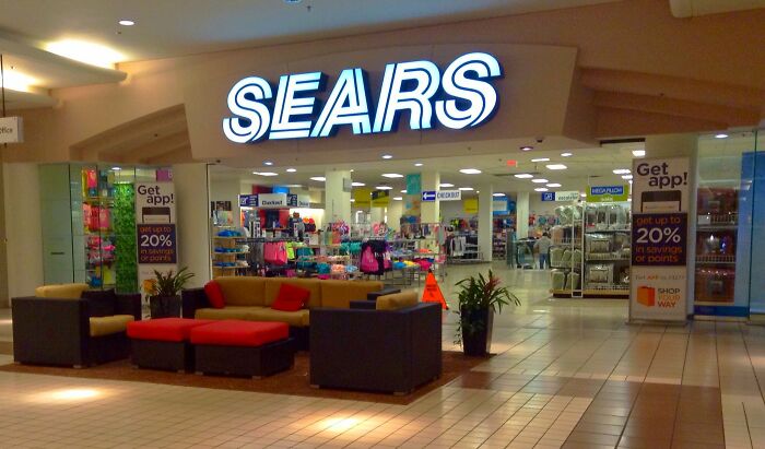 Sears store entrance in a mall with seating area and brightly lit signage visible under surveillance cameras.