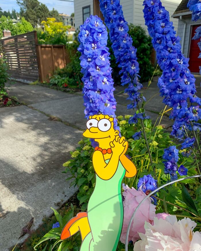 Paper cutout of a cartoon woman interacting with tall blue flowers in a residential garden setting. Paper cutout of a cartoon woman interacting with tall blue flowers in a residential garden setting.