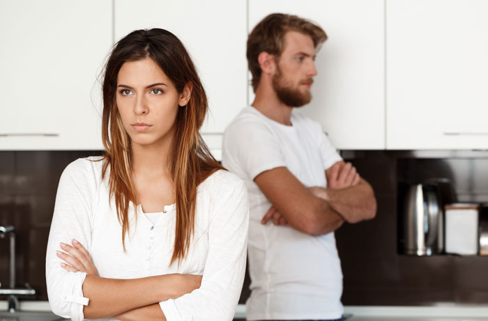 Woman and husband stand apart with arms crossed in kitchen, showing tension after she stops treating him like a kid.
