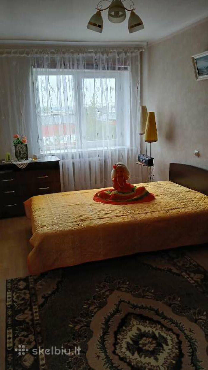 Bedroom in a wild real estate listing with a yellow bedspread, doll centerpiece, floral carpet, and simple furniture by a bright window.