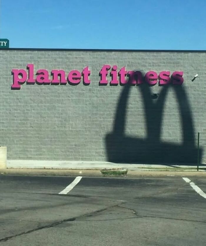 Shadow resembling McDonald's arches cast on a Planet Fitness building wall under a clear blue sky.